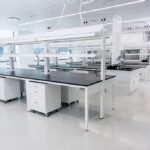 A spacious, modern laboratory with clean white walls and floors, featuring multiple lab benches with black countertops and white drawers on wheels. The lab is well-lit with overhead lighting and large windows. Each bench is equipped with electrical outlets and gas lines hanging from the ceiling, with ample space for equipment and experimentation. The design is open and organized, suggesting a high-functioning, efficient work environment.