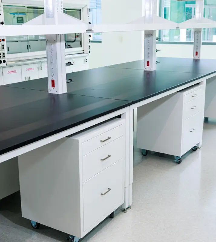 Laboratory benches with black countertops, white drawer units on wheels, and overhead power outlets in a bright lab setting.